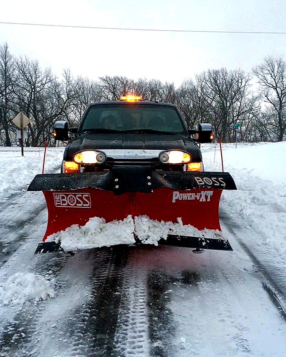 A pickup with a BOSS V-plow pushing snow along a rural road at dusk